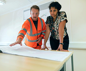 A woman and man review engieering plans spread over table top